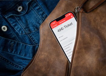 Aus einem geöffneten Reißverschluss einer braunen Handtasche schaut ein Smartphone heraus, auf dem gerade eine Fahrkarten-App mit einem 49€-Ticket darauf geöffnet ist. Im Hintergrund Jeansjacke,